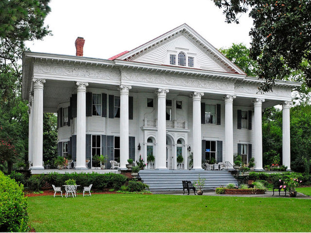 A large white house with huge columns, dark windows, and gray stairs that leads out to a manicured green lawn. 