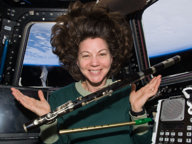 A smiling Cady Coleman in space floating. She wears a green shirt while a clarinet and flute float in front of her. 