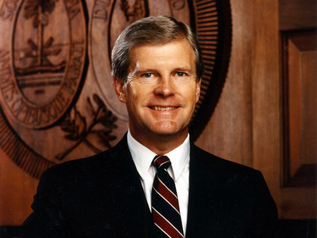 A smiling Carroll Campbell wearing a black suit with a garnet, black and white striped tie. 