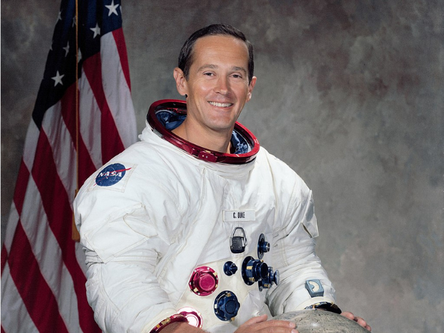 Charles M. Duke in a space suit with a lunar glob in front of him and an American flag behind him. 