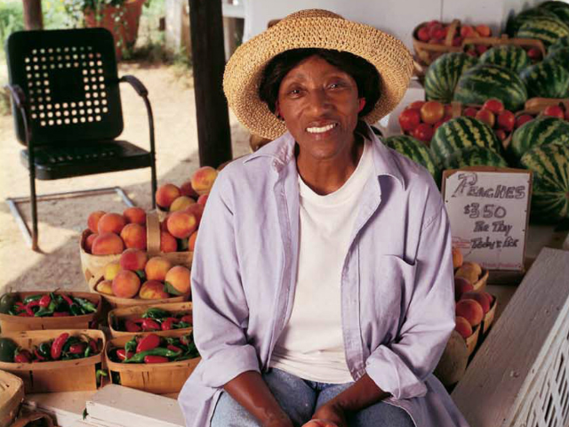 A smiling Dori Sanders wearing a straw hat and lavender shirt over a white t-shit and blue jeans. She sits next to her fruits and vegetables. 