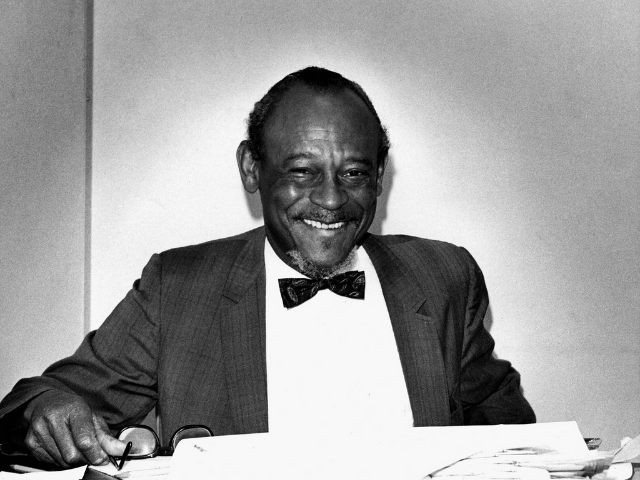 A smiling I. DeQuincey Newman wearing a dark and light suit with a dark bowtie