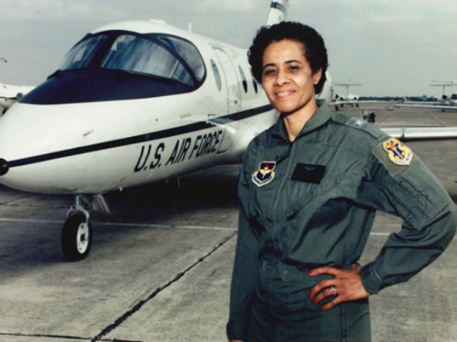 A smiling Irene Trowell-Harris wearing a green air force jumpsuit next to a white plane.