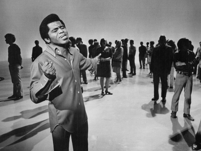 James Brown singing while shadowed people stand in the background 