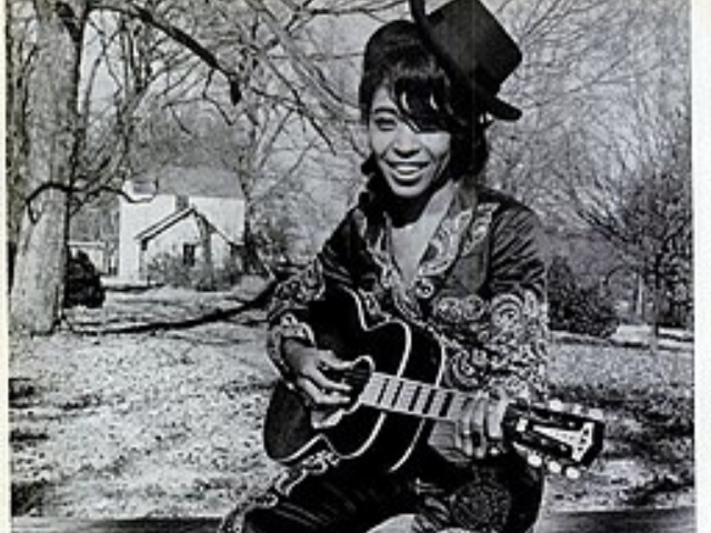 Linda Martell wearing a dark cowboy hat cocked to the side and holding a guitar. 