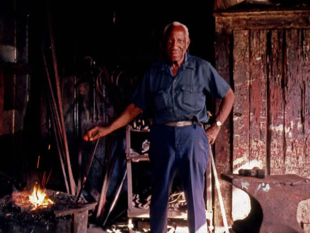 Philip Simmons wearing blue overalls, standing in his forge. 