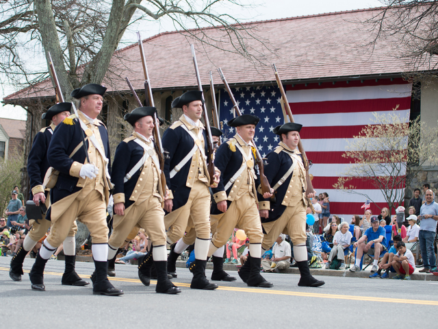 Men dressed in dark blue and tan colonial clothing carrying muskets walking down the street