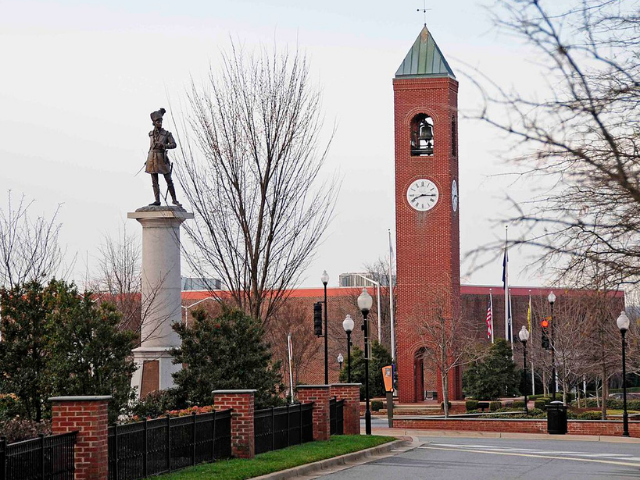 A statue of a man stands on a large white column in the background is a large brick clock tower with a green triangle at the top. 