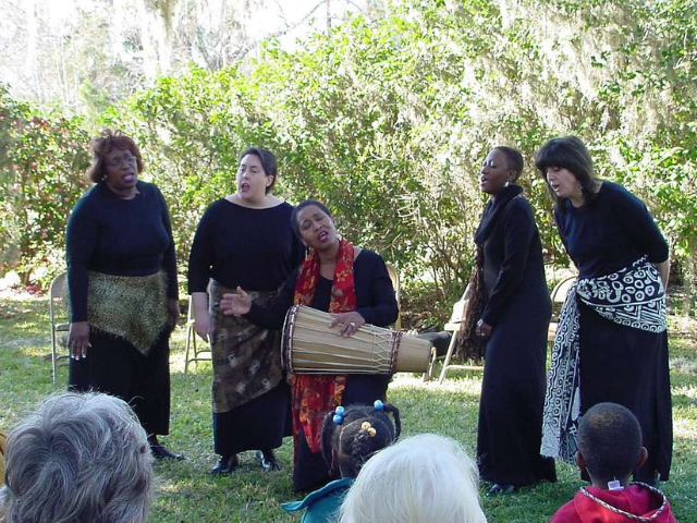 Five women dressed in black and African print sing while one beats on a drum. 