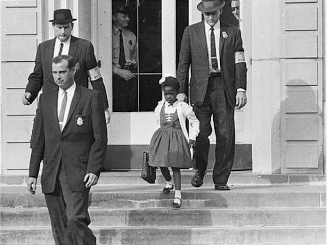 A little girl walks down the stair behind a Man in a dark suit. Two men flank on the sides of the little girl. 