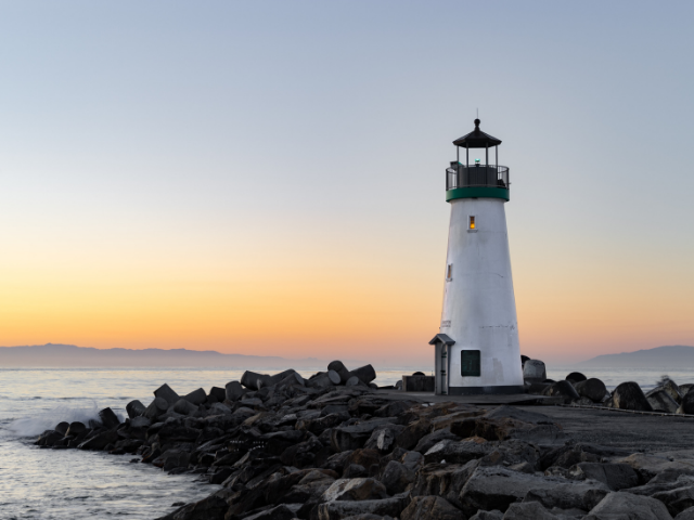 A white lighthouse with a green top on top of dark rocks next to the ocean during the sunrise