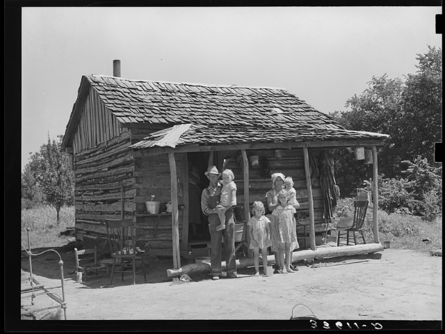 A small wooden house with a man, woman,, and four children standing in front of porch