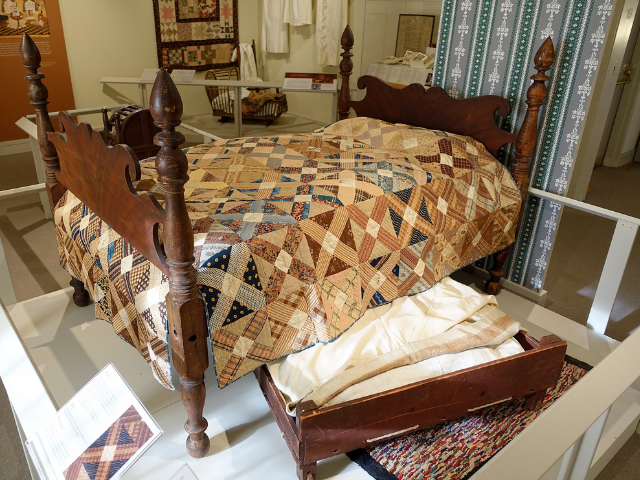 A small bed with a wood frame shows partially under a larger bed with a colorful quilt