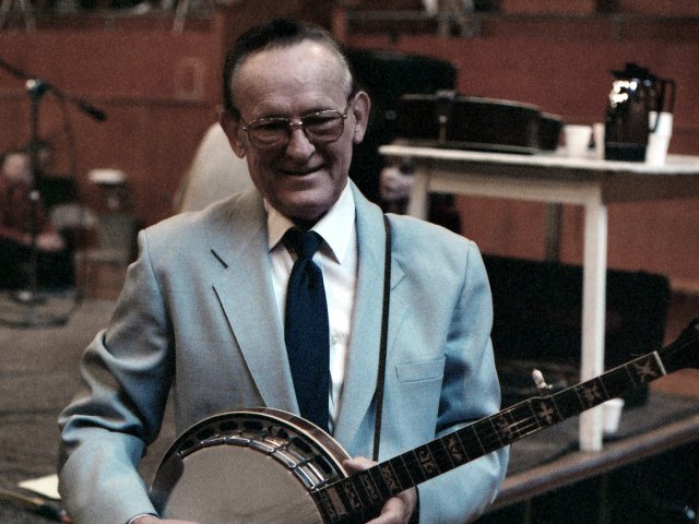 An older man wear a baby blue suit jacket and hold a brown and white banjo.