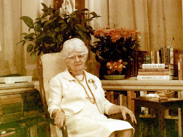 Elderly Laura Bragg sitting in a chair. She wears a white  dress, golden beaded necklace, and broach. 