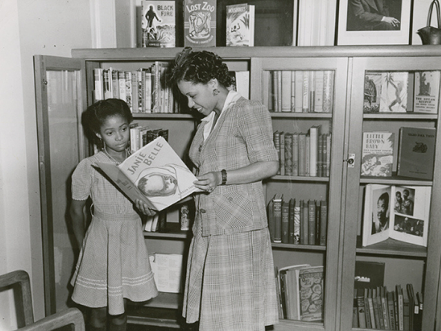 Black and white photograph of Baker showing a book to a child.