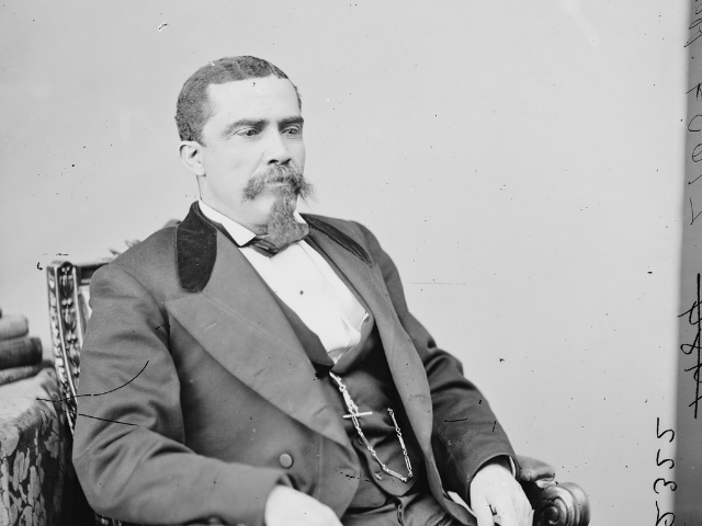An older man in a dark and light suit sitting in a chair.